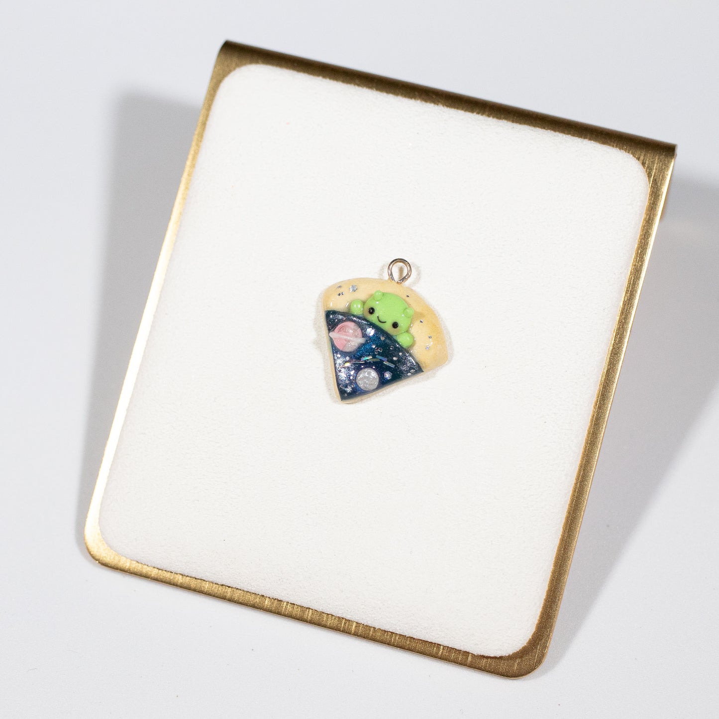 A simple alien pizza charm. This little charm has a tiny, green alien tucked inside a pizza. The pizza is galaxy-themed, with little planets, stars and glitter. This charm can be attached onto a cellphone, planner or purse!