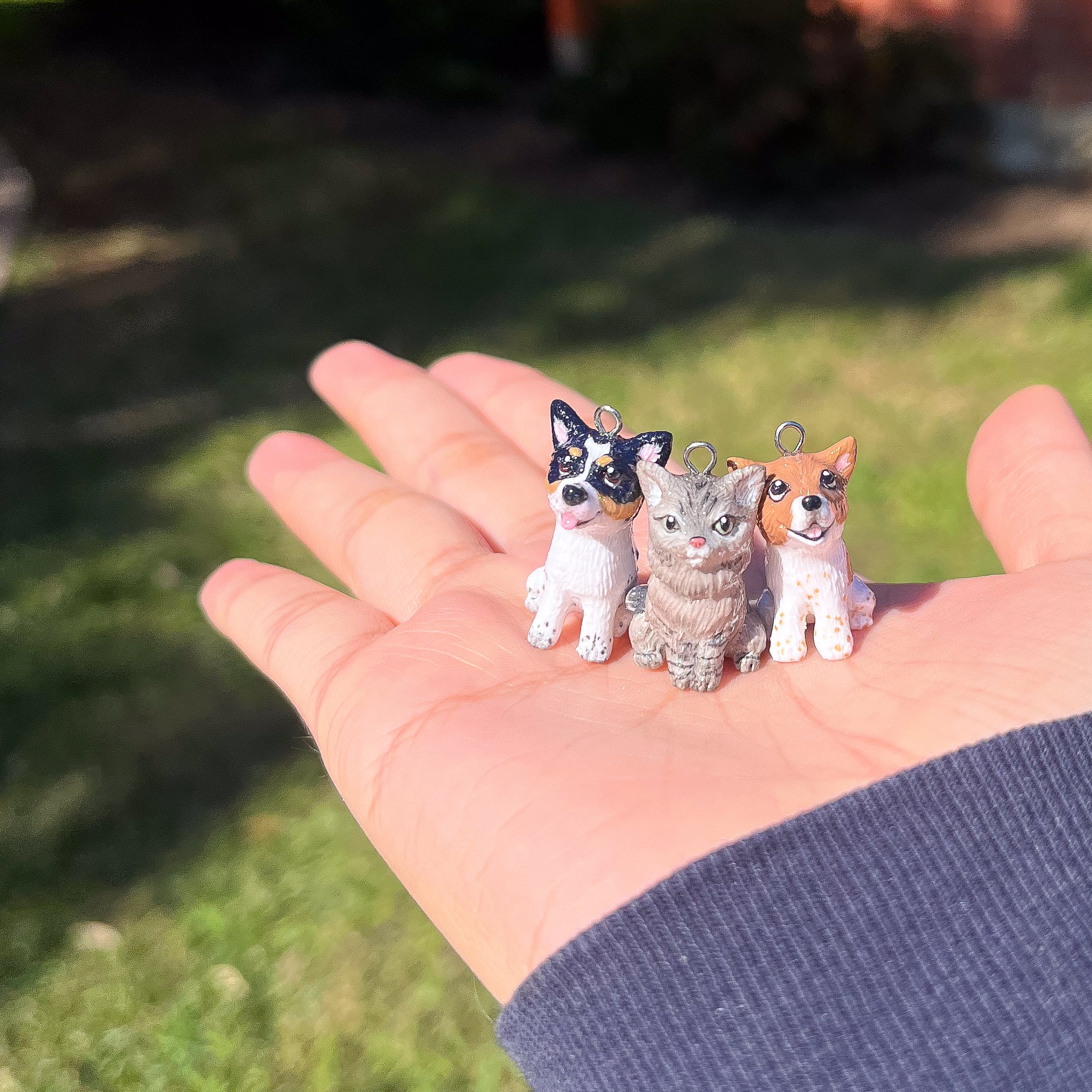 A picture of 3 realistic pet charms in the palm of a hand with an outdoor background. They are about 1 inch in height. There are 2 dog charms and one cat charm. The dogs have brown and black details on their faces and legs. The cat is grey with darker grey markings on it's head and legs.
