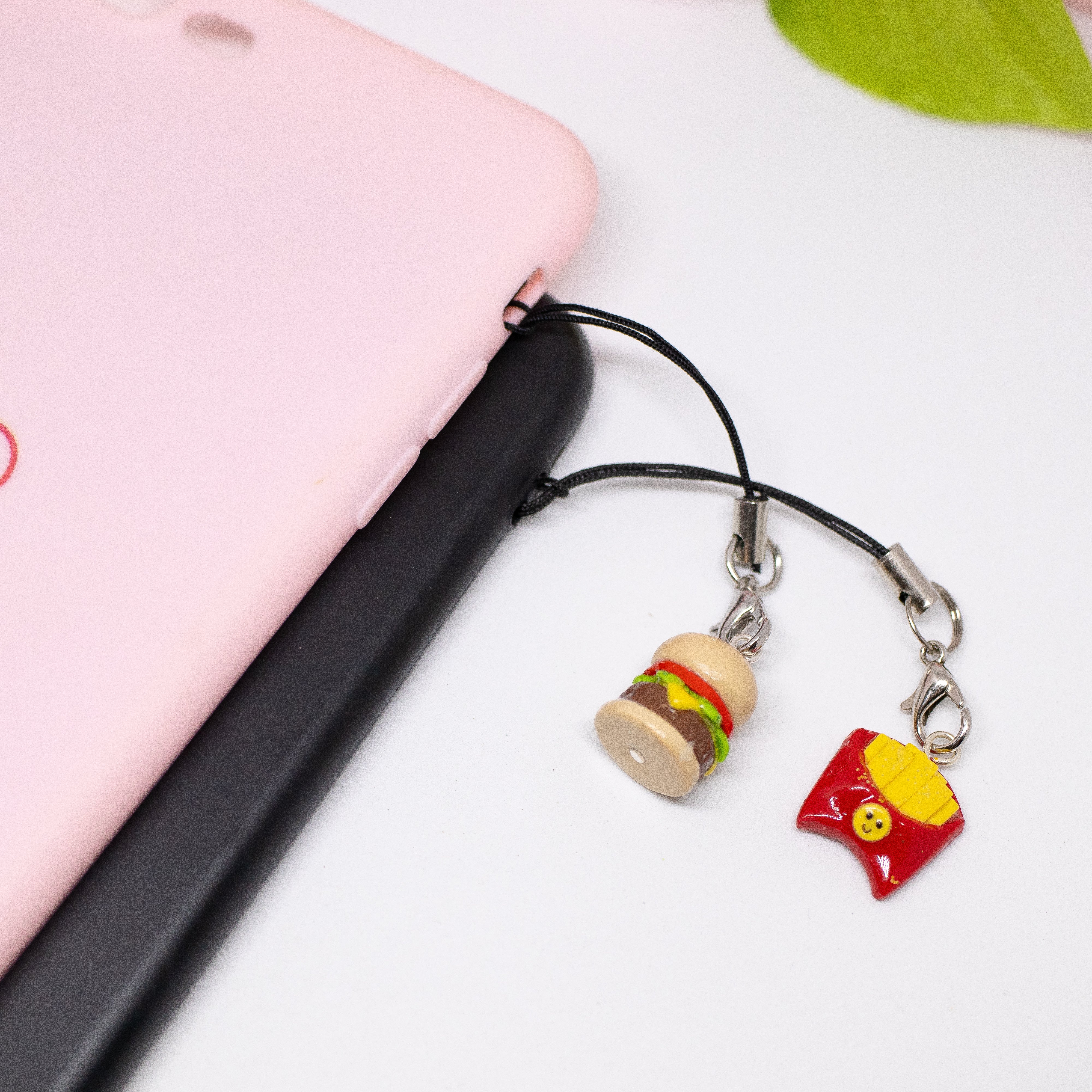 This picture has two phone cases on it against a white background. The first phone case is pink with a little hamburger clay charm attached to it. The second phone case is black with a french fries charm attached to it.