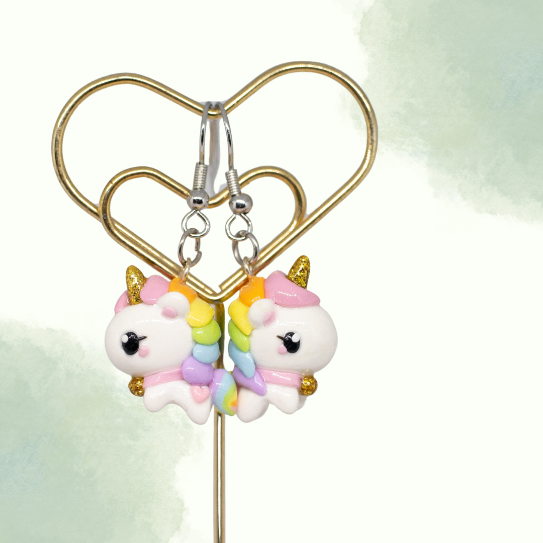 A picture of rainbow unicorn clay earrings hanging from a metal display against a white and green background. 