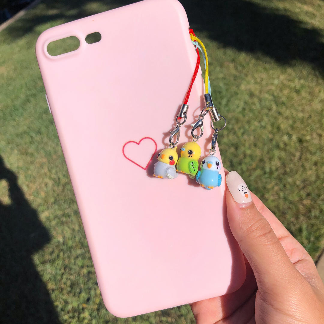 A pink phone case with three different colored budgie charms hanging from the side against a green and black background.