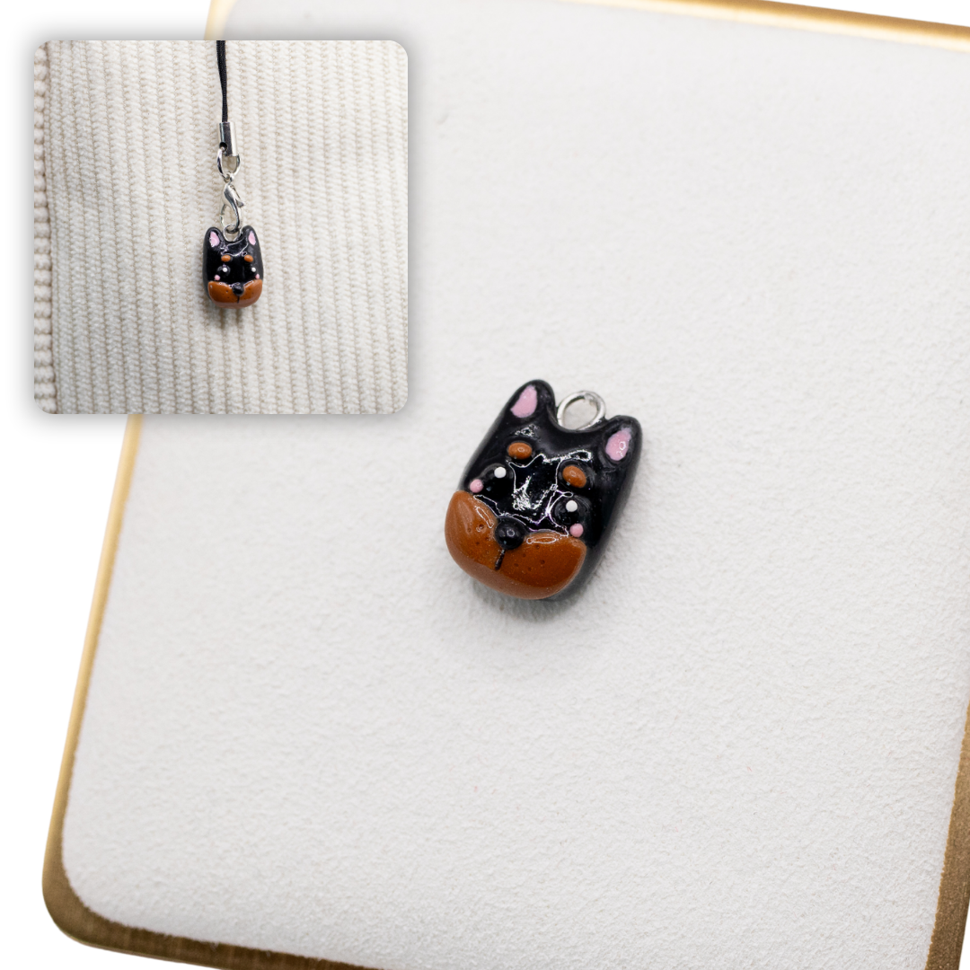 A picture of a dobermann pinscher polymer clay dog charm on a white display with a gold border. There is a 2nd smaller photo of the same dog charm being hung with a phone strap against a textured background.