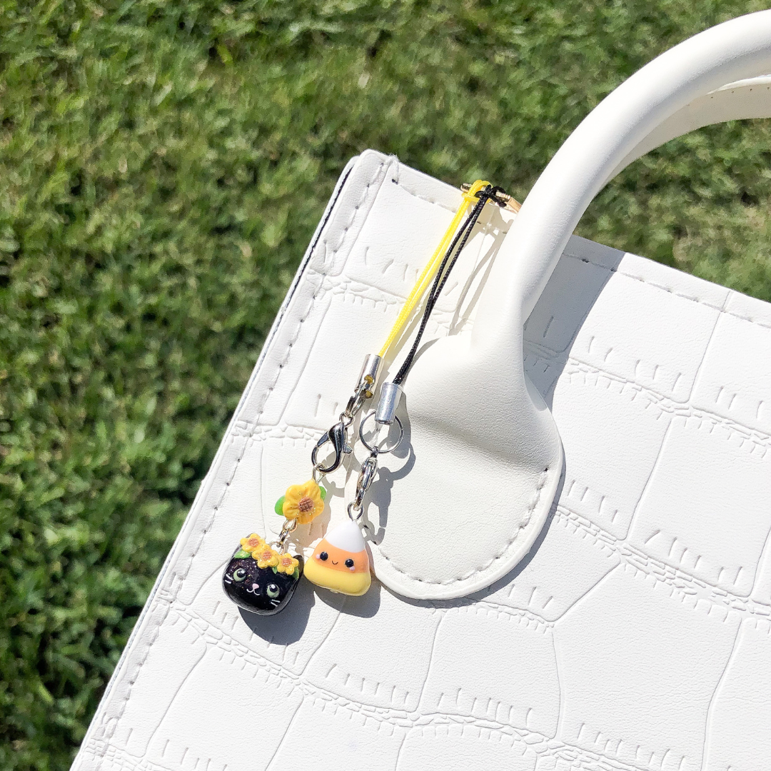 Two fall themed clay charms that are hung on a white purse with a grassy, green background. The first charm is a black cat with sunflowers on it's head and the other is a candycorn charm.