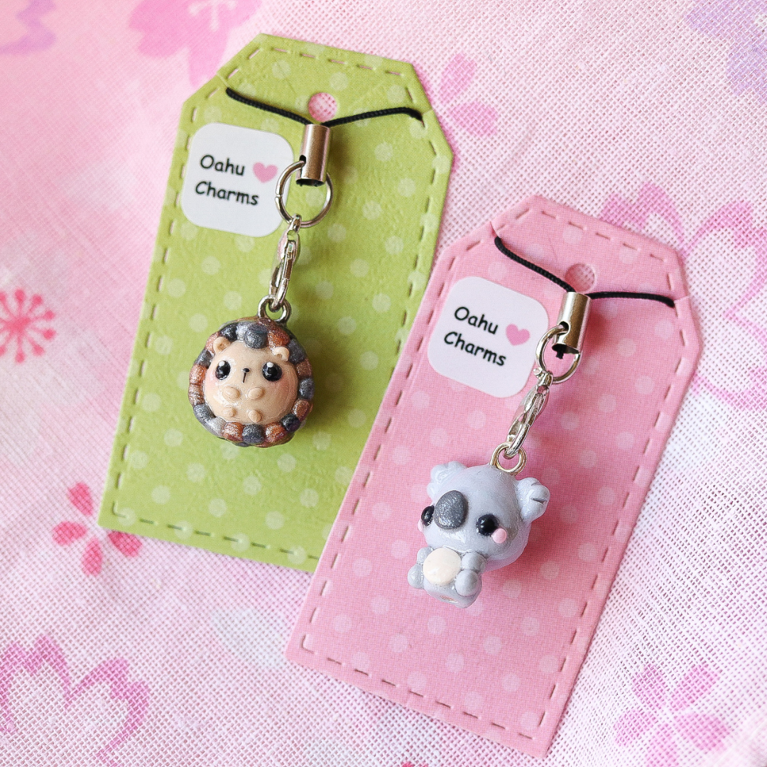 A picture of a hedgehog phone charm with green packaging and a koala phone charm with pink packaging against a pink, floral background.