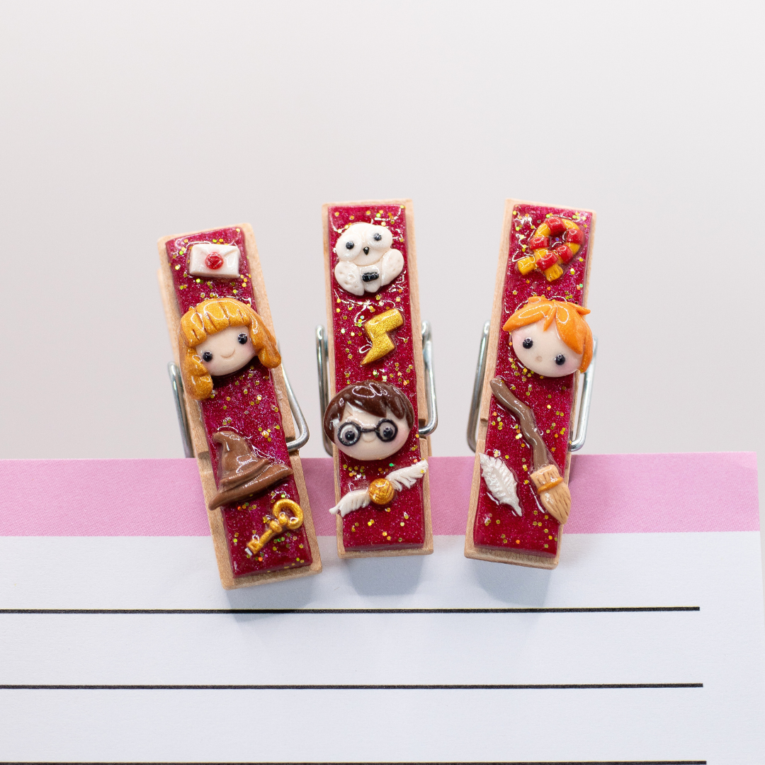 Three decorative clothespins with cute, wizard characters on a white background. The clothespins are clipped onto a notepad for demonstrative purposes.