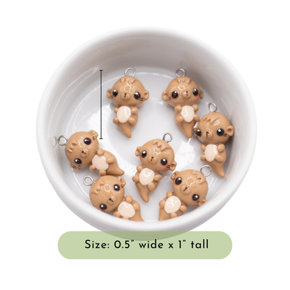 A dish of 7 small otter polymer clay charms held in a white dish with a white background. The picture has text that says, "Size 0.5" wide x 1" tall"