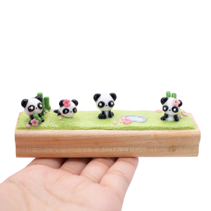 A small figurine of 4 pandas playing together on the grass on a wooden base.
