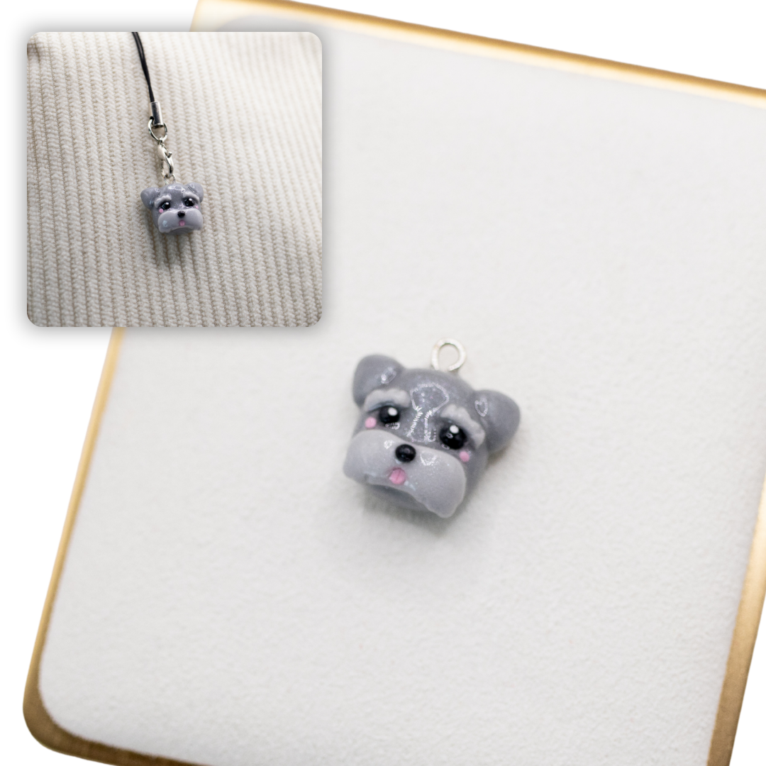 A picture of a grey schnauzer dog polymer clay dog charm on a white display with a gold border. There is a 2nd smaller photo of the same dog charm being hung with a phone strap against a textured background.
