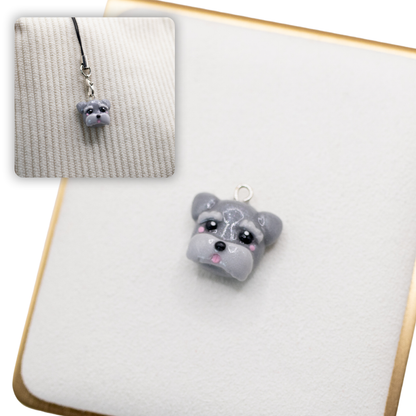 A picture of a grey schnauzer dog polymer clay dog charm on a white display with a gold border. There is a 2nd smaller photo of the same dog charm being hung with a phone strap against a textured background.