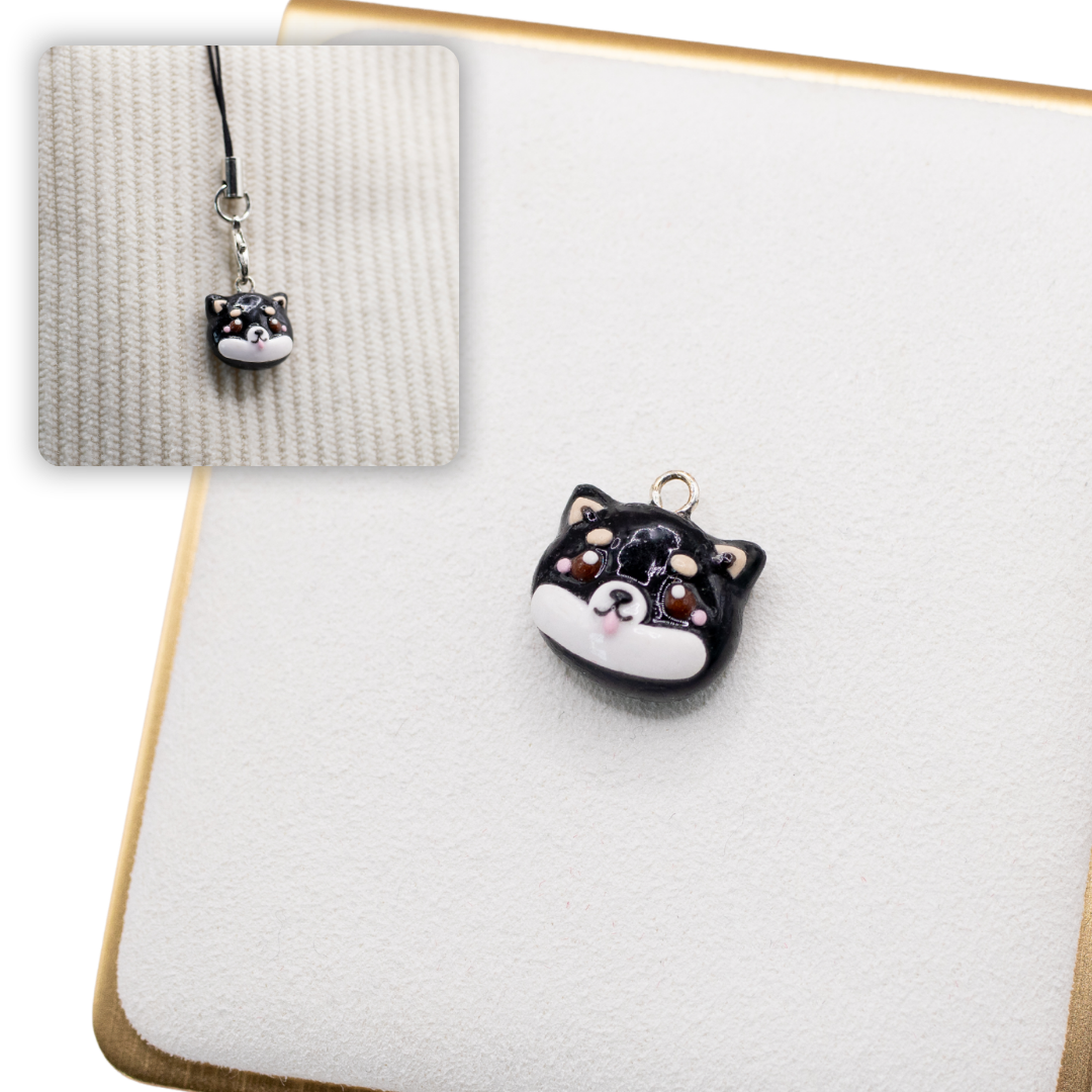 A picture of a black and tan shiba inu polymer clay dog charm on a white display with a gold border. There is a 2nd smaller photo of the same dog charm being hung with a phone strap against a textured background.