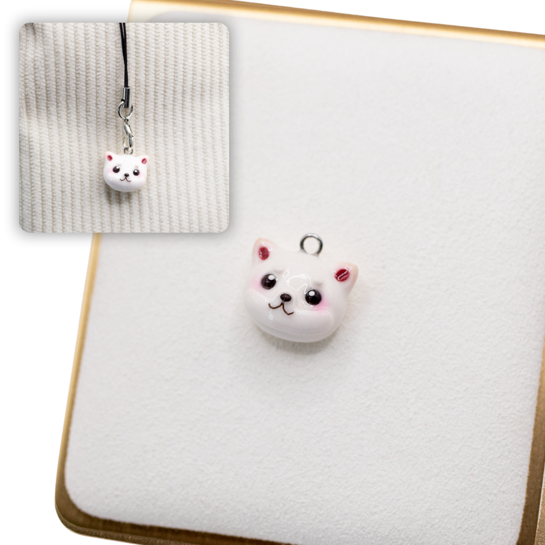 A picture of a cream shiba inu polymer clay dog charm on a white display with a gold border. There is a 2nd smaller photo of the same dog charm being hung with a phone strap against a textured background.