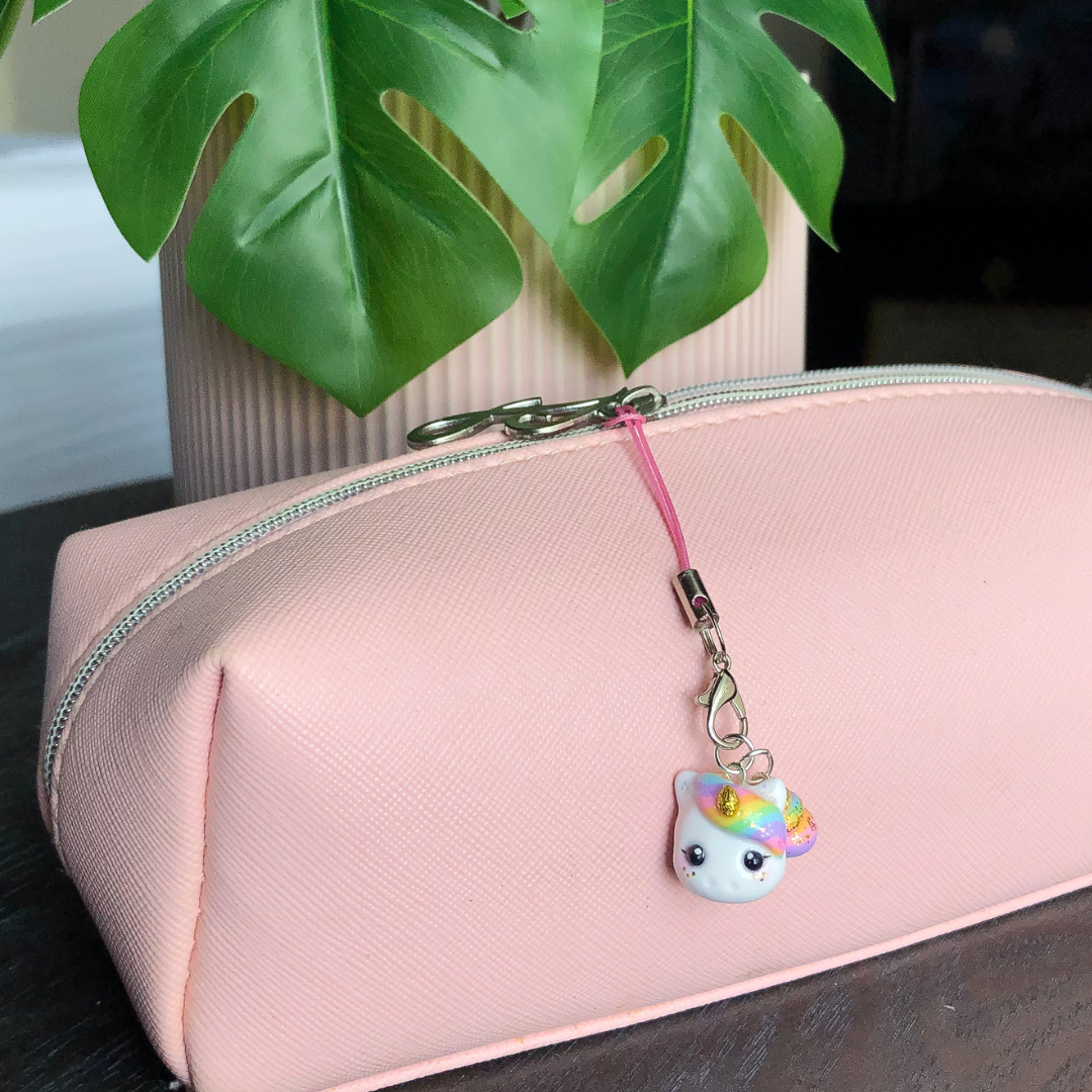 A pink pouch with a unicorn charm hanging from the zipper pull. The background is dark and has a green plant in it.