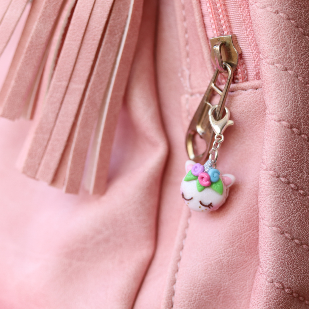 A little unicorn macaron charm used as a zipper pull on a pink backpack. It has three flowers, a silver unicorn horn and a sleepy face with pink blush.